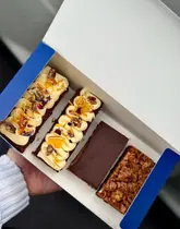 A high-quality, top-down photograph of a bakery box containing four sweet treats: two slices of carrot cake with cream cheese frosting and edible flowers, a chocolate brownie, and a cornflake-topped brownie. The lighting is soft and natural, emphasizing the textures of the baked goods.