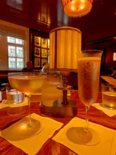 A warm, atmospheric shot of two cocktails/drinks at Simpsons Bar in London, featuring a champagne flute and a coupe glass on a wooden table with a lamp, capturing a sophisticated, historic bar vibe.