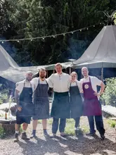 A group portrait of five chefs wearing aprons, standing in a woodland setting in front of large tipis, likely at an outdoor dining event.