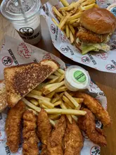 A top-down, casual shot of a meal from Slim Chickens, featuring chicken tenders, Texas toast, fries, a chicken burger, and a milkshake, served on branded paper.