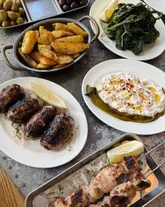A high-quality, overhead flat-lay photograph of a Greek-Cypriot spread featuring various dishes including sheftalia, spicy feta, prawns saganaki, potatoes, and spinach. The lighting is natural and even, highlighting the textures and char marks on the grilled items.
