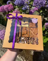 A high-quality, well-composed product shot of a bakery treat box held against a natural, floral background. The lighting is bright and natural, highlighting the textures of the brownies and blondies, while the shallow depth of field keeps the focus entirely on the product.