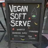 A clear, eye-level photograph of a chalkboard sign placed on a pavement outside a cafe, advertising vegan soft serve ice cream with a list of available flavors.
