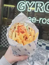 A close-up, handheld shot of a halloumi gyros wrapped in branded paper, held in front of a shop window with neon signage. The focus is sharp on the halloumi and fries, capturing the texture of the street food.