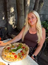 A portrait of a woman holding a Neapolitan-style pizza at an outdoor table. The setting appears to be a casual Italian restaurant with al fresco dining.