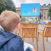 A candid, family-oriented shot capturing a child watching an outdoor screening of 'Bluey' at a public event. The composition uses the child in the foreground to frame the activity, effectively showcasing the event's atmosphere and accessibility for families.