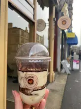 A close-up, eye-level shot of a takeaway tiramisu cup held in a hand, positioned in front of a shopfront. The lighting is natural and even, highlighting the layers of the dessert.