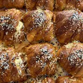 A high-quality, top-down close-up shot of a tray of freshly baked, golden-brown cheese and sesame seed croissants. The image focuses on the flaky texture and generous topping, highlighting the artisan quality of the pastries.