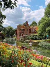 A picturesque, high-quality landscape shot of Westgate Gardens in Canterbury, showcasing a vibrant flower-filled foreground, a river with a punt boat, and a historic brick building in the background under a bright, sunny sky.
