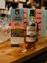 A high-quality, professional product shot featuring a bottle of Loch Lomond 'The Open' Special Edition whisky, its packaging, and a glass of the spirit, set against a blurred background of other bottles in a retail or bar setting.