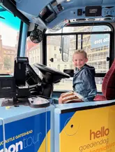 A bright, engaging photograph of a young child sitting in the driver's seat of the Newcastle Toon Tour bus, effectively showcasing the family-friendly nature of the sightseeing activity.