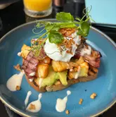 A top-down, close-up shot of a brunch dish featuring a poached egg, bacon, avocado, and halloumi on toast, served on a blue plate.