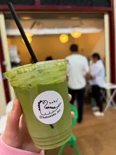 A close-up, first-person shot of a matcha latte in a plastic takeaway cup, held in front of a busy cafe interior.