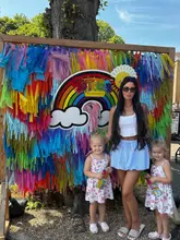 A bright, outdoor photograph featuring a mother and her two young daughters posing in front of a colourful 'TotFest' rainbow backdrop at Windsor Racecourse. The image captures a family-oriented festival atmosphere, with the subjects smiling directly at the camera.