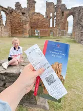 A first-person perspective shot holding a bus ticket and an English Heritage brochure for Tynemouth Priory and Castle, with a child sitting on a stone wall in the background amidst the ruins.