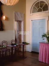 A serene, well-lit interior shot of a cafe space set up for a flower arranging workshop. The image features small tables with floral arrangements, warm lighting, and a pastel-coloured door, creating an inviting and peaceful atmosphere.
