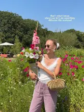 A bright, sunny lifestyle photograph featuring a woman holding a bucket of freshly picked flowers in a field, used to promote a 'Pick Your Own' flower farm experience.
