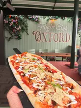 A first-person perspective shot of a long, rectangular pizza being held on a serving paddle at Flight Club Oxford. The background features the venue's branding and outdoor seating area, establishing the location and the casual, social nature of the experience.