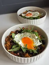 A bright, overhead shot of a homemade brunch dish featuring crispy potatoes, bacon, black pudding, and a fried egg, garnished with fresh rocket. The lighting is natural and even, effectively highlighting the textures of the ingredients.