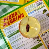 A top-down, flat-lay photograph of a cocktail garnished with a chili, placed directly on top of a Pizza Pilgrims menu. The image captures the vibrant, casual atmosphere of the restaurant, using the menu as a textured background to provide context for the drink.