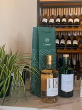A professional product shot featuring two bottles of Llanerch branded spirits and a gift bag, positioned on a counter in front of a wine rack. The lighting is soft and even, highlighting the product labels and the branded packaging effectively.