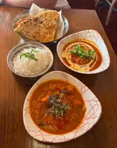A top-down shot of an Indian meal featuring two curries, a bowl of rice, and a basket of naan bread on a wooden table.