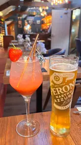 A straightforward, eye-level shot of two drinks—a cocktail garnished with a strawberry and a pint of Peroni beer—placed on a wooden table inside a restaurant setting.