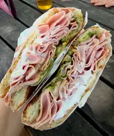 A close-up, handheld shot of a generously filled mortadella and pistachio sandwich, captured outdoors on a wooden table. The image highlights the volume and texture of the ingredients, showcasing the 'stacked' nature of the sandwich.