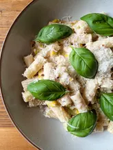 A high-quality, overhead food photograph of a creamy pasta dish featuring sweetcorn, topped with grated parmesan and fresh basil leaves. The lighting is soft and natural, highlighting the textures of the pasta and the freshness of the herbs.