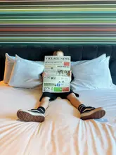 A promotional lifestyle image for Village Hotels featuring a child sitting on a hotel bed holding a 'Village News' information sheet, effectively showcasing the hotel's branding and communication style.
