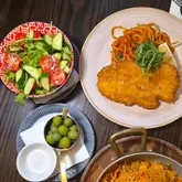 A top-down shot of a lunch spread featuring a breaded chicken dish with pasta, a fresh side salad, olives, and a glimpse of another pasta dish, showcasing a standard Italian restaurant meal.