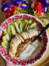 A bright, overhead shot of a healthy breakfast yogurt bowl featuring sliced banana, kiwi, and granola. The composition is clean and aesthetically pleasing, utilizing natural light and floral elements to create an inviting, fresh look.