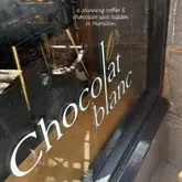 A static shot of a shop window for 'Chocolat Blanc' in Hamilton. The image features the shop's name on the glass and a text overlay, but the composition is heavily impacted by reflections of the street, which obscures the interior and makes the image feel cluttered.