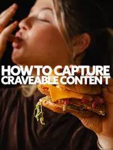 A high-quality, dark and moody close-up photograph of a person holding a burger, used as an example of 'craveable' food content. The image focuses on the texture and appeal of the food, utilizing shallow depth of field to isolate the subject.