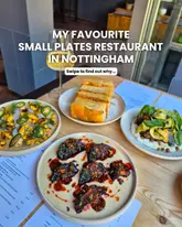 A bright, overhead flat-lay shot of a small plates spread featuring focaccia, roasted beetroot, charred courgettes, and a corn-based dish, accompanied by a glass of white wine. The image is well-lit with natural light and clearly showcases the variety and presentation of the food.