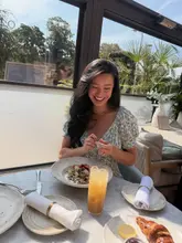 A bright, lifestyle-focused shot of a woman enjoying breakfast on a terrace, featuring a granola bowl, a croissant, and a cocktail. The image captures a relaxed, premium dining atmosphere, though the production quality is slightly marred by visible filming equipment in the background.