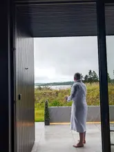 A lifestyle shot featuring a person in a bathrobe standing in the doorway of a modern suite, holding a coffee mug and looking out at a view of a loch. The image aims to convey a sense of relaxation and luxury, highlighting the scenic location and the comfort of the accommodation.