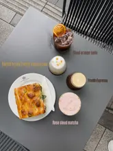 A clean, well-composed flat-lay photograph showcasing a selection of four distinct drinks and a focaccia sandwich on a grey outdoor table. The image uses natural lighting to highlight the textures of the food and the vibrant colours of the beverages, effectively promoting the cafe's menu.