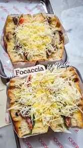 The video provides a quick tour of a focaccia sandwich shop, showing the exterior, the counter display, the menu, and close-ups of the prepared sandwiches topped with parmesan. It uses a handheld style with music and text overlays to present the venue as a casual spot for a quick bite.