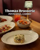 A professionally captured, atmospheric shot of a crab crumpet at Thomas Brasserie in Cardiff. The image uses a shallow depth of field to isolate the dish, creating a warm, inviting restaurant ambiance.