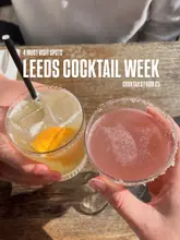 A POV shot of two people holding cocktails over a wooden table, promoting Leeds Cocktail Week.