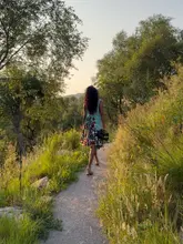 A scenic, lifestyle-focused travel shot featuring a person walking along a path in the Scottish Highlands during golden hour. The image captures the natural beauty of the landscape, including trees, tall grass, and the soft, warm light of the setting sun, promoting a tranquil and picturesque getaway experience.