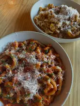 An overhead shot of two bowls of pasta served on a wooden table, featuring a beef ragu pappardelle and a tomato-based pasta, both topped with grated cheese.