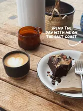 A high-angle, natural light shot of a coffee and a brownie on a rustic wooden table, likely taken at a cafe during a day trip. The image features a latte with heart art and a brownie with a bite taken out of it.