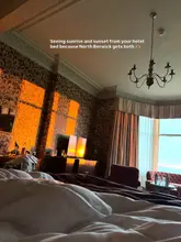 A high-quality, atmospheric shot of a hotel room interior at The Marine Hotel in North Berwick, capturing the warm, golden light of sunrise or sunset streaming through the window. The composition effectively highlights the room's comfort and the scenic coastal view, creating an inviting and aspirational travel aesthetic.