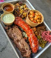 A high-angle, close-up shot of a BBQ platter featuring brisket, sausage, pulled pork, beans, coleslaw, and various sauces, served on a metal tray.