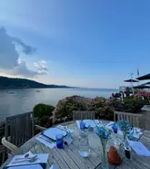 A high-quality, wide-angle shot of an outdoor dining table set for a meal at a restaurant overlooking the sea. The image captures the serene atmosphere of a coastal venue, with the table in the foreground and a beautiful sunset view over the water in the background.