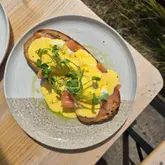 A top-down shot of an Eggs Royale dish served on a ceramic plate, placed on a wooden table outdoors. The dish features poached eggs, smoked salmon, and hollandaise sauce garnished with pea shoots on toasted sourdough.