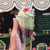 A close-up shot of a matcha soft serve ice cream cup held in a hand, featuring distinct layers of matcha ice cream and red bean paste, topped with a decorative flower. The background shows a green-themed pop-up stall with a menu board, creating a cohesive and on-brand aesthetic for a Japanese-inspired dessert spot.