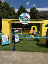 A photo of the 'Stay & Play' event entrance in Old Eldon Square, Newcastle. The image shows a bright yellow archway with branding, leading into an outdoor play area with artificial grass, featuring various play structures and families enjoying the space.